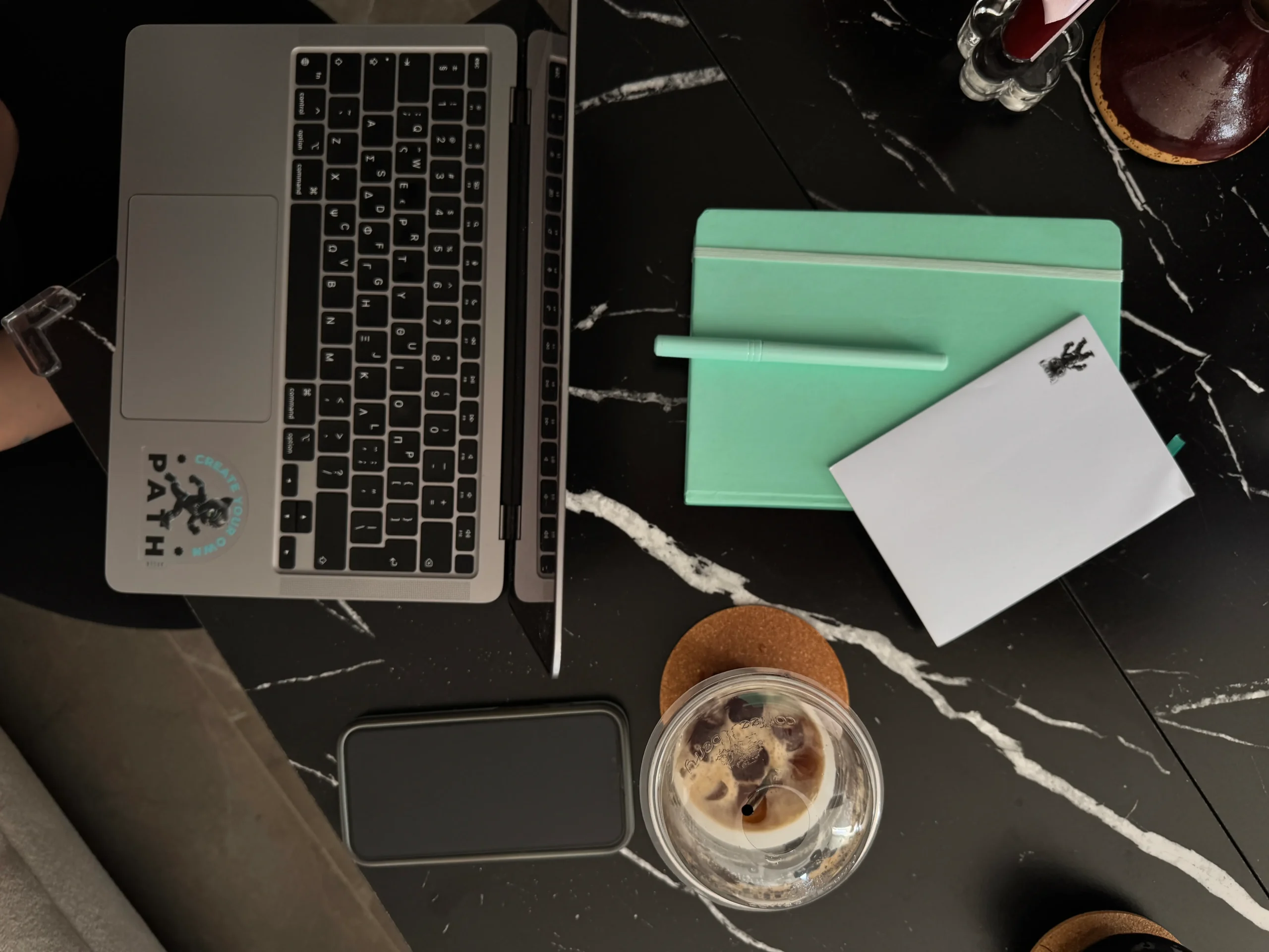 Overhead view of a workspace with a MacBook laptop, mint green notebook, notepad, phone, and iced coffee on a black marble surface