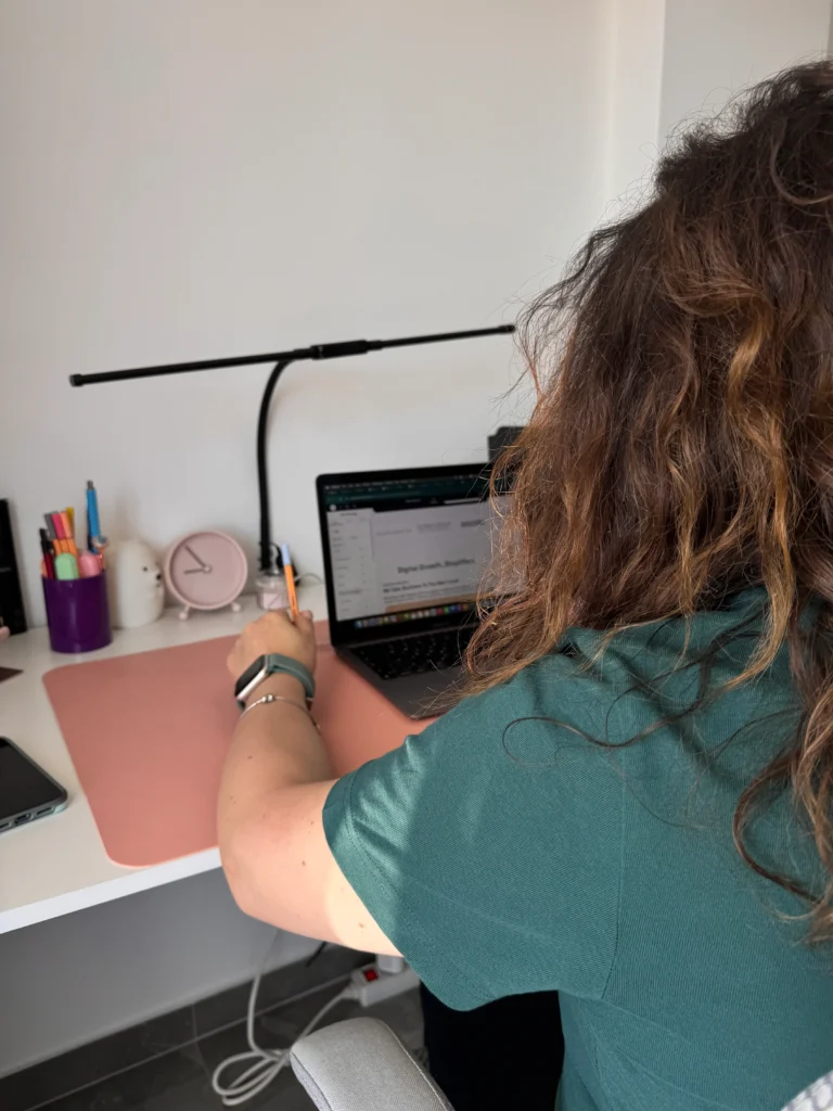 Woman with curly hair seen from behind, working at a desk on a laptop showing a website, holding a pen with colourful stationery and a pink clock nearby