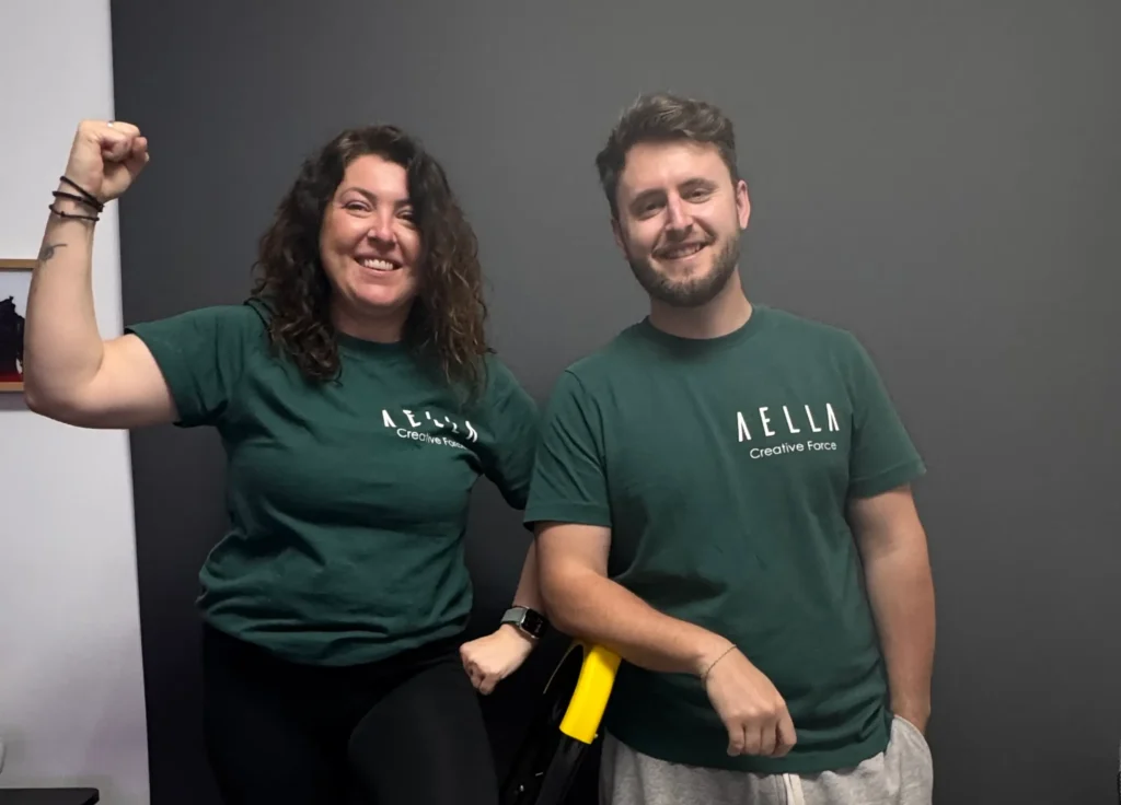 Two smiling team members wearing dark green Aella Creative Force branded t-shirts, one with fist raised in celebration, against a grey studio wall