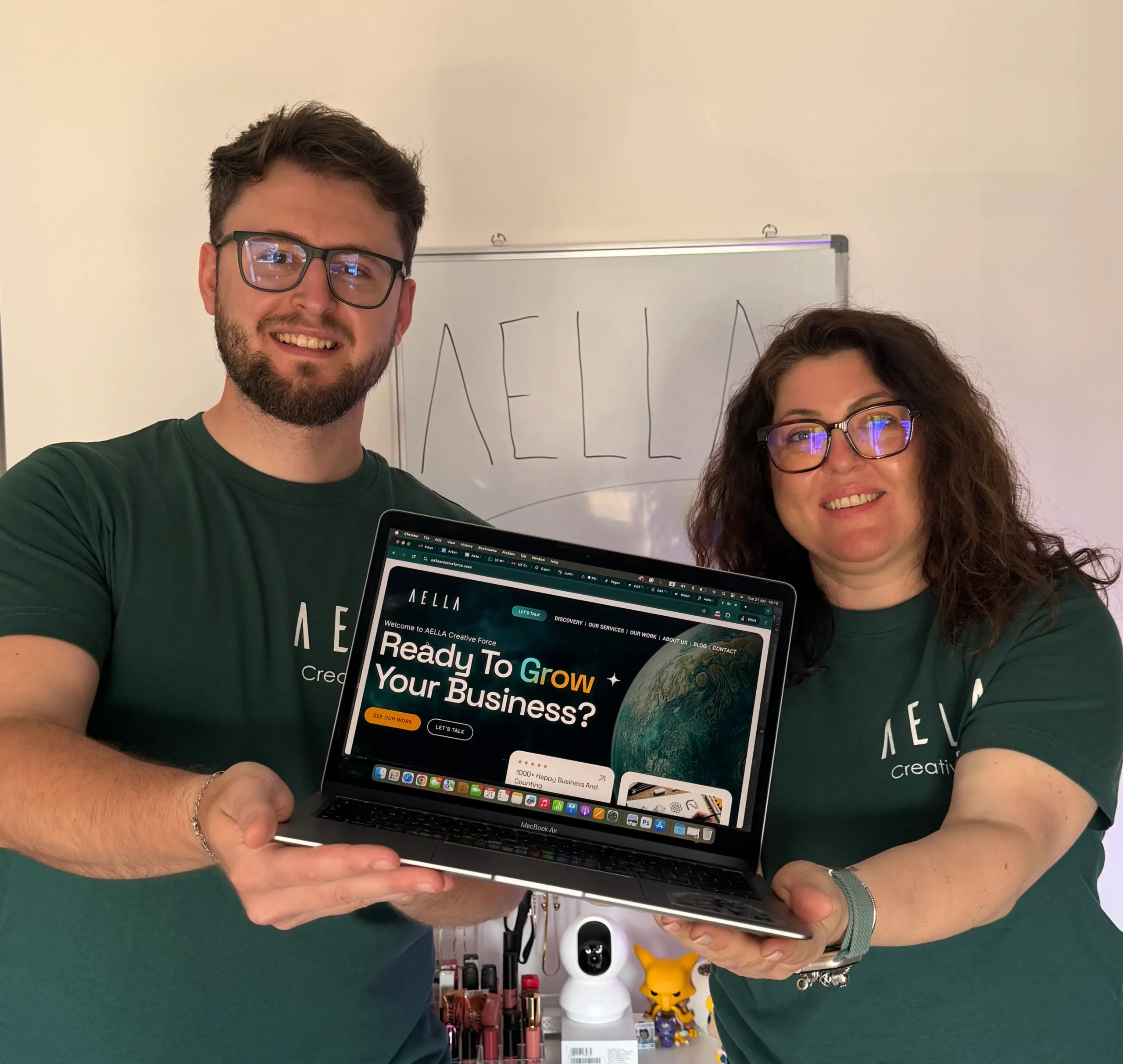 Two co-founders of Aella Creative Force, Kiki and Briken, in dark green branded t-shirts smiling and holding a MacBook displaying the Aella website, with the Aella logo written on a whiteboard behind them