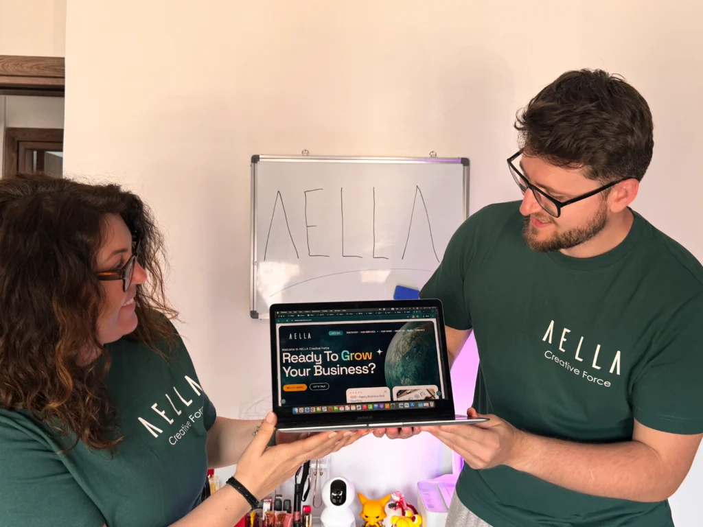 Two Aella Creative Force team members in dark green branded t-shirts looking at and discussing the Aella website on a MacBook, with the Aella logo on a whiteboard behind them